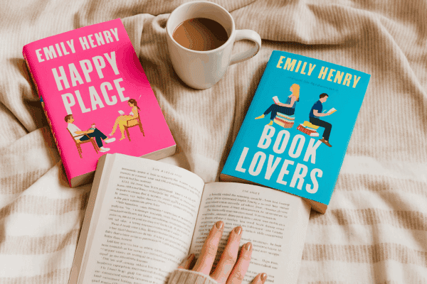 A cozy reading nook featuring Emily Henry’s Happy Place and Book Lovers, with an open book in hand, a mug of coffee, and a striped beige blanket.